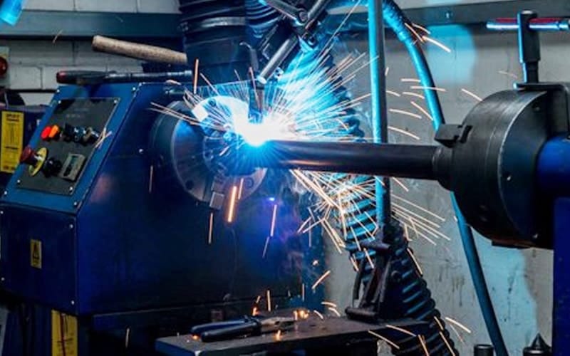 Robotic welding arm creating bright sparks while welding an industrial shaft in an automated manufacturing facility.